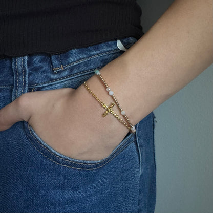 A close-up of a person's wrist wearing two delicate bracelets: one gold chain with a cross charm and one gold chain with small white beads, styled with blue jeans and a black top.