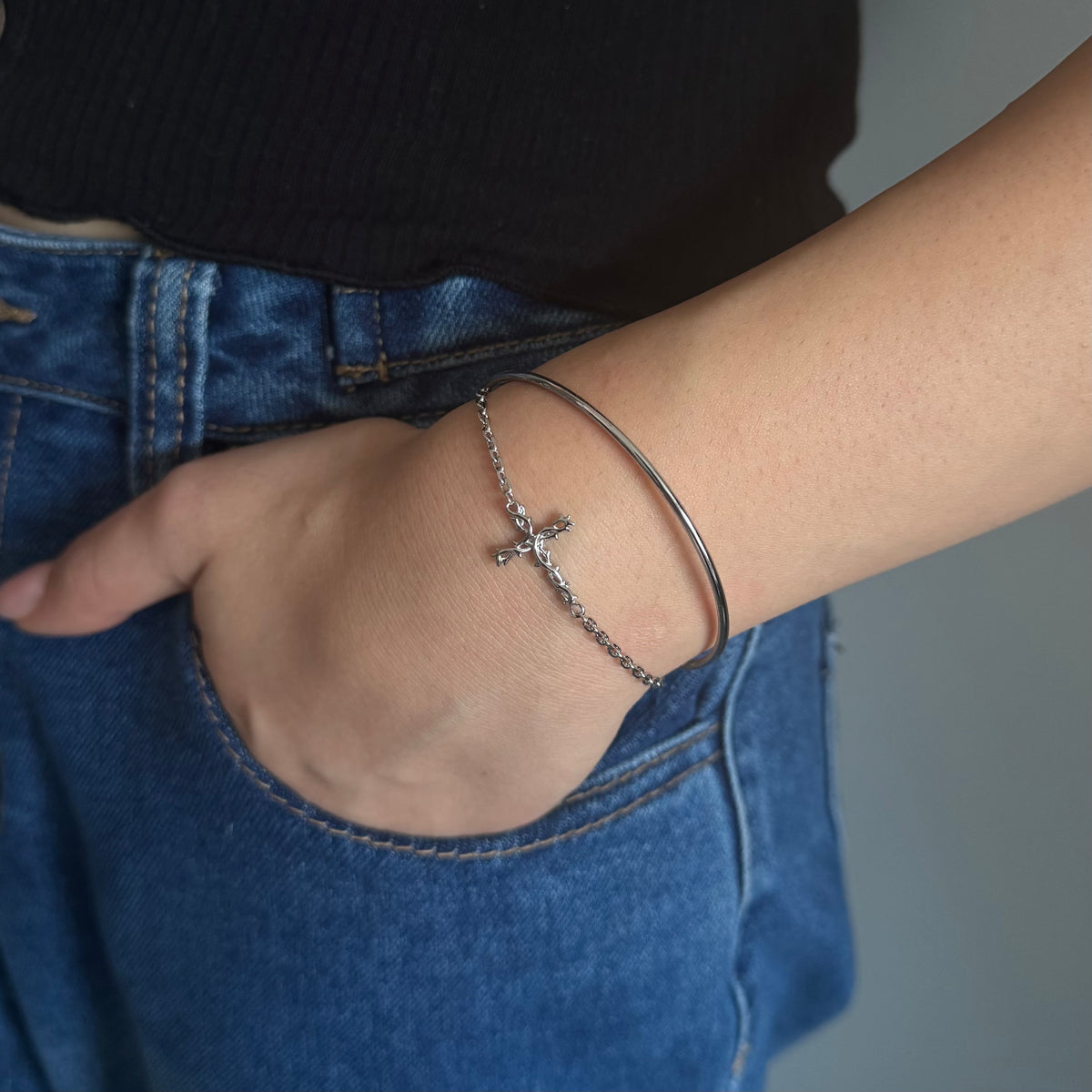 A close-up of a person's wrist wearing a silver chain cross bracelet and a thin silver bangle, paired with blue jeans and a black top.