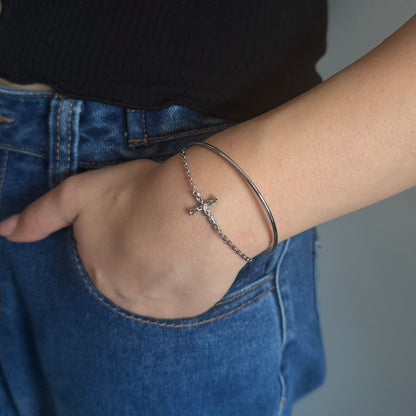 A close-up of a person's wrist wearing a silver chain cross bracelet and a thin silver bangle, paired with blue jeans and a black top.
