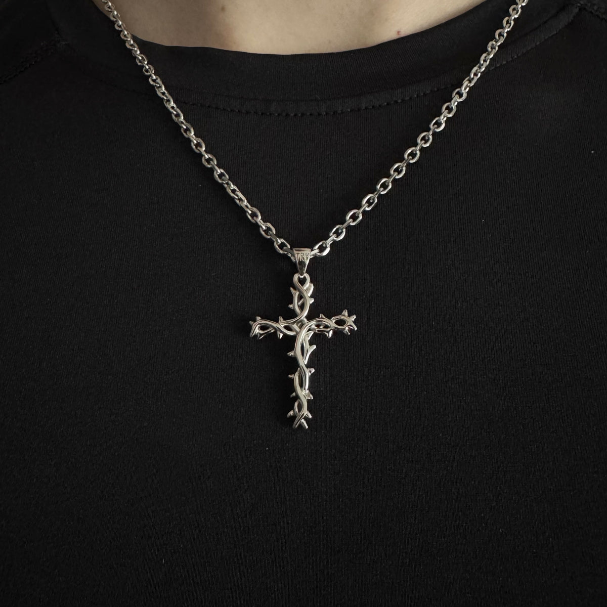A silver cross pendant necklace with a thorny design, worn on a chain over a black t-shirt.