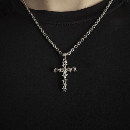 A silver cross pendant necklace with a thorny design, worn on a chain over a black t-shirt.